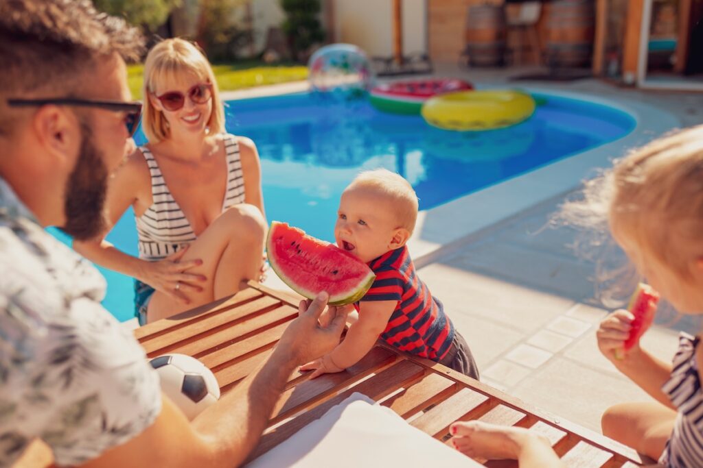 Parents having fun spending hot sunny summer day by the swimming pool with their children, father feeding baby boy with a slice of watermelon