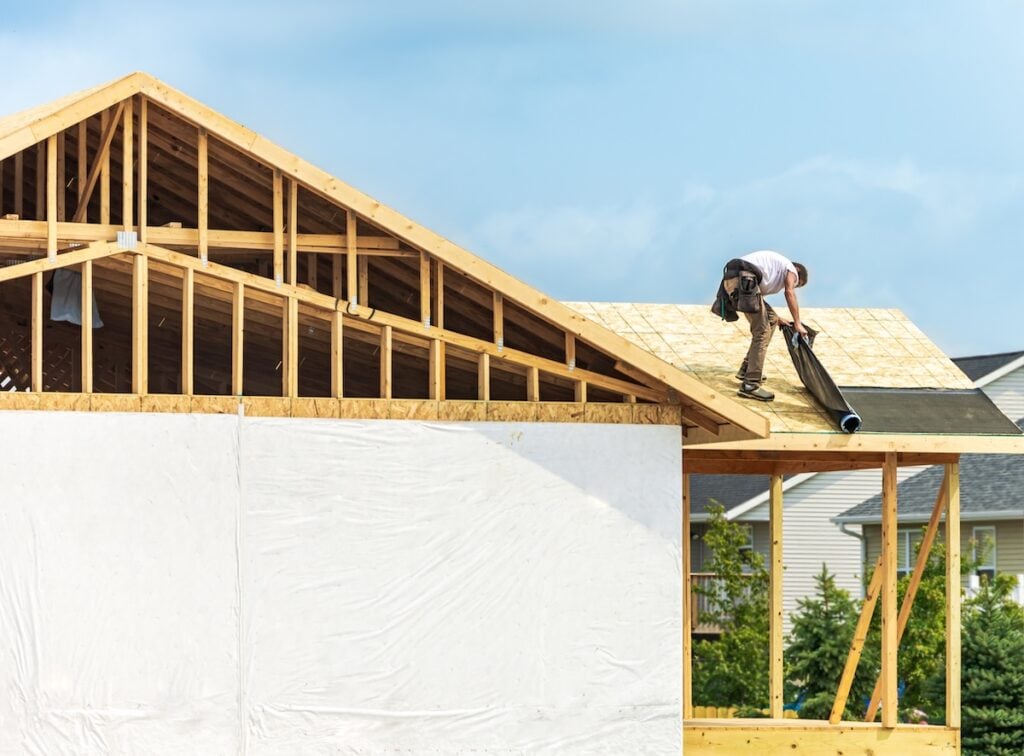 Worker installing underlayment sheet on wooden roof of new house. 